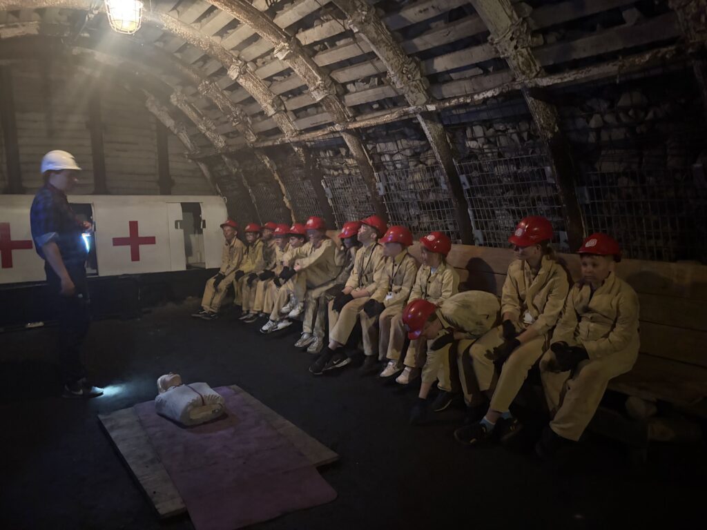 Grupa dzieci w strojach sztygarskich i czerwonych kaskach siedzi na ławce w podziemiach i słucha opowieści przewodnika.