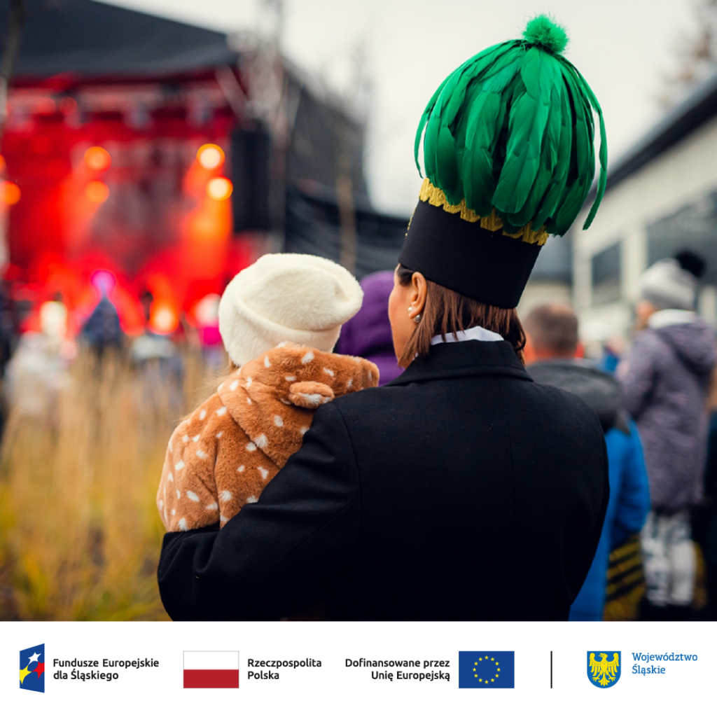 Kobieta w galowym stroju górniczym i czako z zielonymi piórami stoi odwrócona tyłem, trzyma na rękach dziecko i patrzy w kierunku sceny